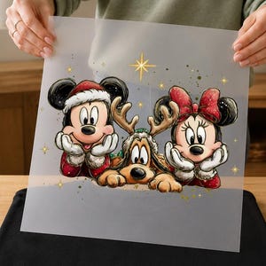 May include: A square, semi-transparent transfer sheet featuring a festive illustration of Mickey Mouse in a Santa hat, Minnie Mouse with a red bow, and Pluto wearing reindeer antlers, all accented with gold stars and glittery speckles.
