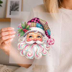 May include: A square transparent sheet featuring a colorful illustration of Santa Claus with a swirling white and pink beard, wearing a patchwork hat with pink plaid, leopard print, and green holly accents, topped with a pink and white striped pom-pom.