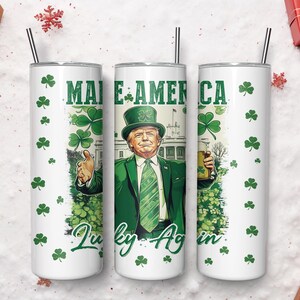 May include: White tumbler with a green and white design featuring a cartoon of a man in a green suit and hat, with the text "Make America Lucky Again" and shamrocks. The tumbler has a black straw.