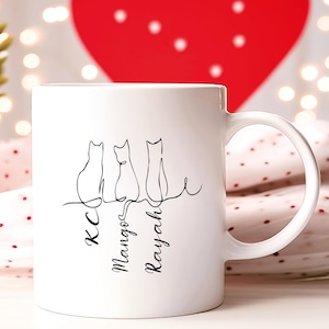 May include: White ceramic mug with a black line drawing of three cats sitting on a line. The names "KC", "Mangos", and "Rayah" are written underneath the cats.