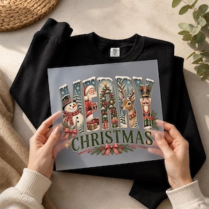 May include: A black crewneck sweatshirt featuring a transparent transfer sheet with the text MERRY CHRISTMAS. The letters are filled with holiday imagery including a snowman, Santa Claus, a decorated Christmas tree, a reindeer, and a nutcracker.