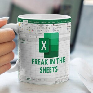 K&ouml;nnte beinhalten: Eine wei&szlig;e Kaffeetasse mit einem gr&uuml;nen Microsoft Excel-Tabellenblatt-Design. Die Tabelle zeigt den Text "FREAK IN THE SHEETS".