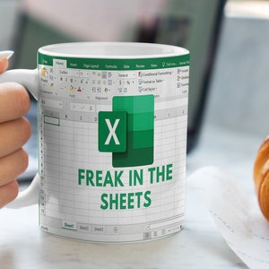 K&ouml;nnte beinhalten: Wei&szlig;e Kaffeetasse mit einem gr&uuml;nen und wei&szlig;en Microsoft Excel-Tabellenblatt-Design. Die Tabelle zeigt den Text "FREAK IN THE SHEETS" an.