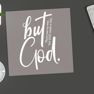 May include: A square sign with a taupe background featuring white text that reads but God. There was no way, but GOD made a way. The sign is positioned on a dark gray surface alongside a white keyboard, a black pen, and a small green plant.