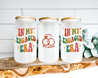 May include: Three frosted glass tumblers with bamboo lids and straws. The tumblers have different designs. One says "In My Engaged Era" in colorful retro lettering. Another says "Future Mrs. Johnson" with a heart and two rings. The third tumbler has the same design as the first.