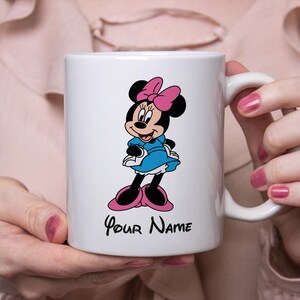 Puede incluir: Taza de cer&aacute;mica blanca con un dise&ntilde;o de dibujos animados de Minnie Mouse. Minnie lleva un vestido azul, un lazo rosa y zapatos. La taza tiene el texto "Your Name" en escritura negra debajo del personaje.
