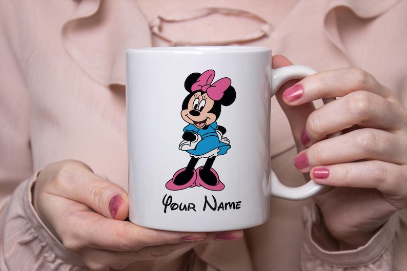 Puede incluir: Taza de cer&aacute;mica blanca con un dise&ntilde;o de dibujos animados de Minnie Mouse. Minnie lleva un vestido azul, un lazo rosa y zapatos. La taza tiene el texto "Your Name" en escritura negra debajo del personaje.