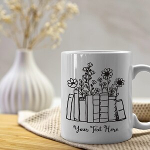 Puede incluir: Taza de cer&aacute;mica blanca con un dibujo lineal negro de libros y flores. El texto "Your Text Here" est&aacute; debajo de los libros. La taza tiene un asa curva. La taza est&aacute; sobre una superficie de madera y un pa&ntilde;o beige.