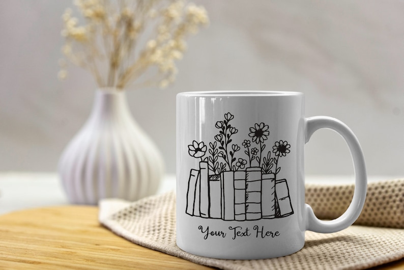 Puede incluir: Taza de cer&aacute;mica blanca con un dibujo lineal negro de libros y flores. El texto "Your Text Here" est&aacute; debajo de los libros. La taza tiene un asa curva. La taza est&aacute; sobre una superficie de madera y un pa&ntilde;o beige.