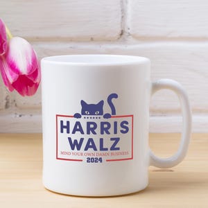 May include: White ceramic coffee mug with a blue and red graphic of a cat peeking over a sign that says "Harris Walz Mind Your Own Damn Business 2024".