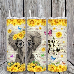 May include: A stainless steel tumbler with a design of a cartoon elephant wearing glasses and a sunflower crown. The background is a white brick wall with sunflowers and butterflies. The text "You are my sunshine" is printed on the tumbler.