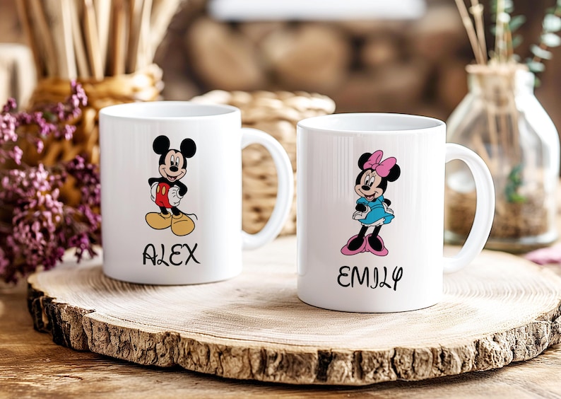 Puede incluir: Dos tazas de cer&aacute;mica blanca con ilustraciones de Mickey y Minnie Mouse. Mickey lleva pantalones cortos rojos y zapatos amarillos, Minnie un vestido azul y zapatos rosas. Las tazas tienen los nombres "ALEX" y "EMILY".