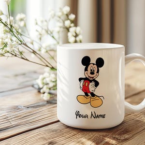 Puede incluir: Taza de cer&aacute;mica blanca con una ilustraci&oacute;n de Mickey Mouse. El personaje lleva pantalones cortos rojos, zapatos amarillos y tiene orejas negras. El texto "Your Name" est&aacute; impreso debajo. La taza tiene un asa curvada.