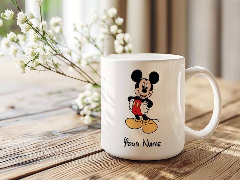 Puede incluir: Taza de cer&aacute;mica blanca con una ilustraci&oacute;n de Mickey Mouse. El personaje lleva pantalones cortos rojos, zapatos amarillos y tiene orejas negras. El texto "Your Name" est&aacute; impreso debajo. La taza tiene un asa curvada.