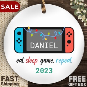 May include: White round Christmas ornament with a colorful illustration of a Nintendo Switch video game console with the text "DANIEL" on the screen. The ornament also includes the text "eat. sleep. game. repeat. 2023".
