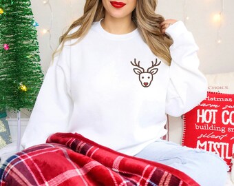 May include: White sweatshirt with a small brown reindeer graphic on the chest. The reindeer has antlers and a heart-shaped nose. The sweatshirt is part of a Christmas-themed collection.