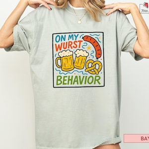 Puede incluir: Camiseta verde claro con un gráfico colorido que dice "ON MY WURST BEHAVIOR". El gráfico incluye dos jarras de cerveza, una salchicha y un pretzel. La camiseta es de corte holgado.