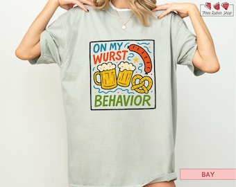 May include: A light green t-shirt with a colorful graphic that reads "ON MY WURST BEHAVIOR." The graphic includes two mugs of beer, a sausage, and a pretzel. The shirt is a relaxed fit.