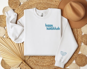 May include: White sweatshirt with the text "happy hanukkah" in blue script on the chest. A blue menorah design is on the left sleeve. The sweatshirt is displayed on a woven surface with a hat in the background.
