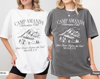May include: White and pepper-colored t-shirts with a mountain and camping graphic. The text on the shirts reads "CAMP AMANDA Adventure Club" and "Last Trail Before the Veil MOAB, UT". The shirts are short-sleeved.