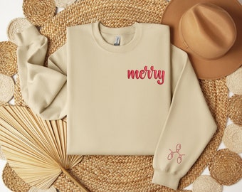 May include: A beige sweatshirt with the word "merry" embroidered in red on the chest and a string of red Christmas lights on the sleeve. The sweatshirt is displayed on a woven surface with a brown hat.