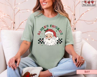 May include: Sage green t-shirt with the text "Santa's Favorite" above a cartoon Santa Claus face. The Santa illustration features a red hat, white beard, and a smiling expression. A black and white checkered pattern is below the Santa illustration.