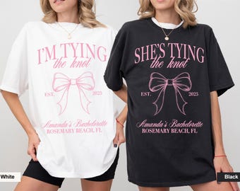 May include: Two white and black t-shirts with the text "I'm Tying the Knot" and "She's Tying the Knot" in pink. Both shirts have a pink bow with the text "EST. 2025" and "Amanda's Bachelorette Rosemary Beach, FL".