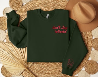 May include: Dark green sweatshirt with the embroidered phrase "don't stop believin'" in pink. The sleeve features a pink embroidered Santa Claus design. The sweatshirt is displayed on a woven surface with a brown hat in the background.