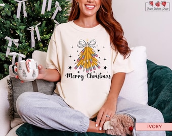 May include: Ivory-colored t-shirt with a Christmas tree design made of pencils, a polka-dot bow, and the words "Merry Christmas." The shirt is a relaxed fit and is paired with gray sweatpants. A mug with Christmas trees is held in the hand.