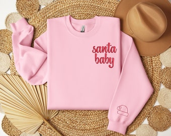 May include: A light pink sweatshirt with the text "santa baby" in red script on the chest. A small red Santa hat is embroidered on the left sleeve. The sweatshirt is displayed on a woven surface with a brown hat in the background.