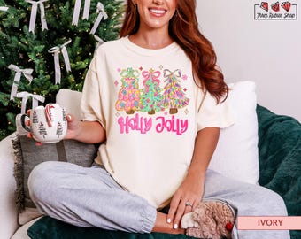 May include: Ivory-colored t-shirt with a colorful graphic of three Christmas trees and the words "Holly Jolly" in pink. The trees are decorated with bows and ornaments. The shirt is worn by a person.