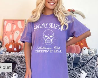 May include: Violet-colored t-shirt with the text "Spooky Season" above a skull graphic. Below the skull, the text reads "Halloween Club CREEPIN' IT REAL". The shirt is part of a Halloween-themed collection.
