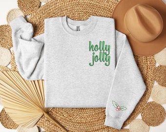 May include: A light gray sweatshirt with the words "holly jolly" embroidered in green on the chest. The sleeve features a small embroidered holly and berry design. The sweatshirt is a festive holiday apparel item.