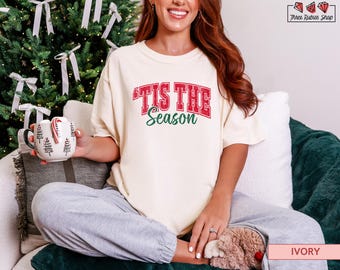 May include: Ivory-colored t-shirt with the text "Tis the Season" in red and green. The model is holding a mug with Christmas tree designs and candy canes. The shirt is paired with gray sweatpants.