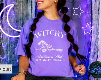 May include: A violet t-shirt with the word "WITCHY" above a witch on a broom graphic. Below the graphic, the text reads "Halloween Club BREWING UP SOME MAGIC". The shirt is part of a Halloween-themed collection.