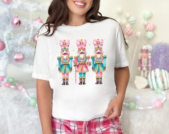 May include: White t-shirt with a graphic of three nutcracker soldiers wearing pink and blue uniforms with pink bows on their hats. The nutcrackers are holding a candy cane, a present, and a candy cane.