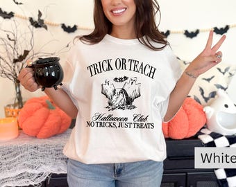 May include: White t-shirt with the text "TRICK OR TEACH" and a Halloween-themed graphic. The shirt also has the text "Halloween Club NO TRICKS, JUST TREATS". A black cauldron mug is held in the image. The shirt is a Halloween-themed product.