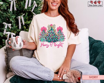 May include: Ivory-colored t-shirt with a Christmas tree design and the text "Merry & Bright" in pink script. The graphic features three decorated trees with pink bows and a black and white polka dot bow. A mug with Christmas tree designs is held in the left hand.