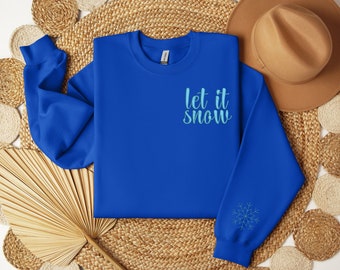 May include: A royal blue sweatshirt with the text "Let it snow" in light blue script on the chest. A snowflake design is embroidered on the sleeve. The sweatshirt is folded and displayed on a woven surface.