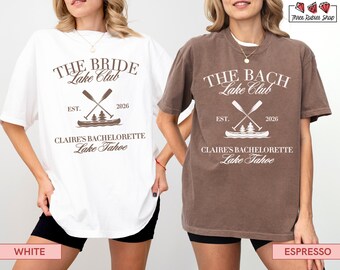 May include: Two oversized t-shirts, one white and one espresso, featuring the text "THE BRIDE Lake Club" and "THE BACH Lake Club" respectively. Both shirts include a canoe graphic with crossed oars, the year 2026, and "Claire's Bachelorette Lake Tahoe".