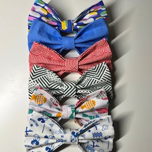 May include: Five colourful bow ties with different patterns. The top bow tie is blue with a yellow and pink pattern. The second bow tie is red with a white pattern. The third bow tie is green and white with a geometric pattern. The fourth bow tie is white with a yellow and red fish pattern. The bottom bow tie is white with a blue Mickey Mouse pattern.