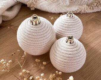 May include: Three cream-colored, rope-wrapped Christmas ornaments with gold tops. The ornaments are on a wooden surface with dried white flowers.