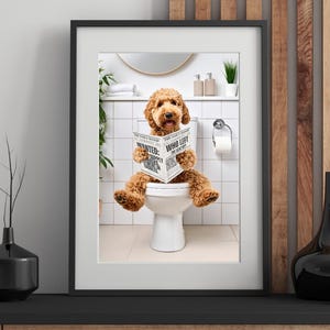 May include: A framed print featuring a brown Goldendoodle dog sitting on a toilet and reading a newspaper. The newspaper headline reads "WANTED: SUPPLY WHO LEFT THE SEAT UP". The bathroom has white tile and a mirror.