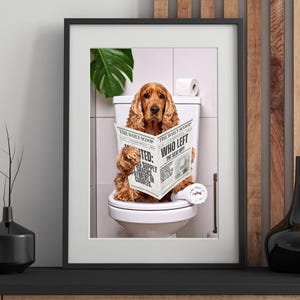 Op de afbeelding: Een ingelijste print van een Cocker Spaniel op een toilet, lezend in een krant getiteld "The Daily Scoop". De hond heeft bruin, krullend haar. De krantenkop luidt "Who Left The Seat Up?" Een rol toiletpapier is zichtbaar.