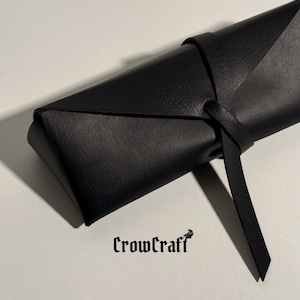 Op de afbeelding: Een zwarte leren schede met een striksluiting. De schede is gevouwen en gestikt, met een riem die om het lichaam wikkelt en aan de zijkant vastknoopt. De tekst "CrowCraft" is rechtsonder in het zwart gedrukt.