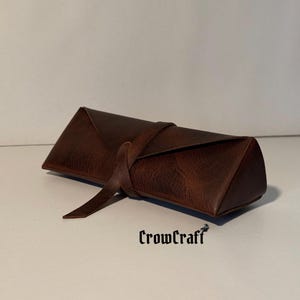 Op de afbeelding: Een bruine leren oprolbare etui met een driehoekige vorm. De etui is vastgezet met een bijpassende leren riem. De tekst "CrowCraft" is zichtbaar op het oppervlak.