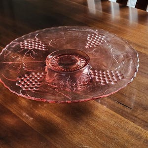 May include: A pink glass cake stand with a grape design. The stand has a scalloped edge and a raised center.