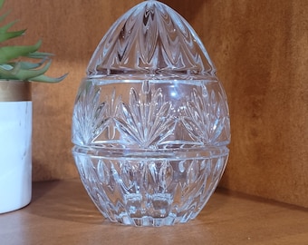 May include: A clear, cut-glass egg-shaped container with a lid, displayed on a wooden shelf. The container is made of clear glass with a detailed, patterned design. The lid and base are separate pieces, and the container is empty.