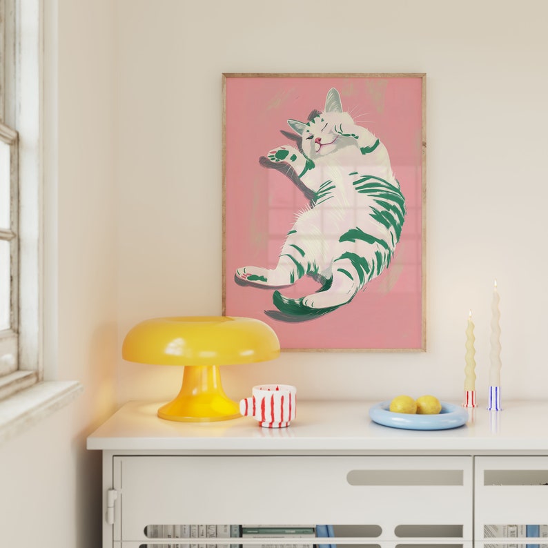 May include: A framed illustration of a white cat with green stripes on a pink background. The cat is lying on its back with its paws in the air. The frame is made of wood and has a light brown finish.