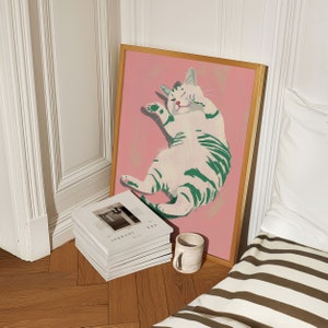 May include: A framed illustration of a white cat with green stripes lying on its back on a pink background.